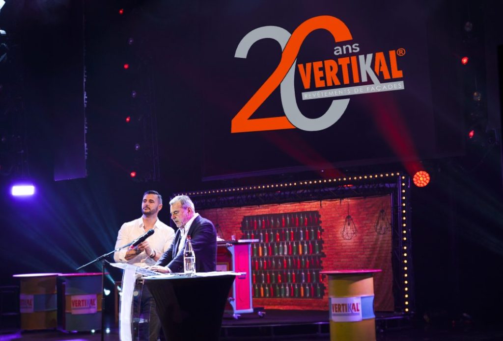 Deux hommes sur scène avec un logo et un message pour les 20 ans VERTIKAL.