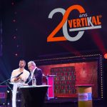 Deux hommes sur scène avec un logo et un message pour les 20 ans VERTIKAL.
